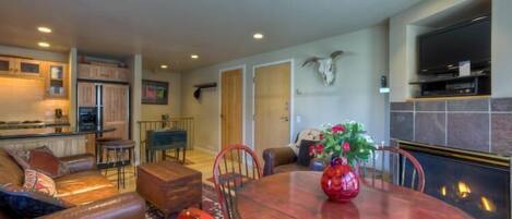 A spacious living area with a dining table, comfortable seating, a kitchen in the background, and a mounted TV.