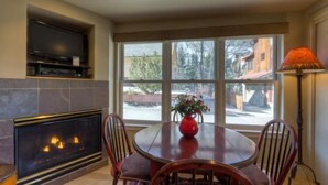 A room with a fireplace, a dining table with chairs, a television mounted on the wall, and a large window with a view of the outside.