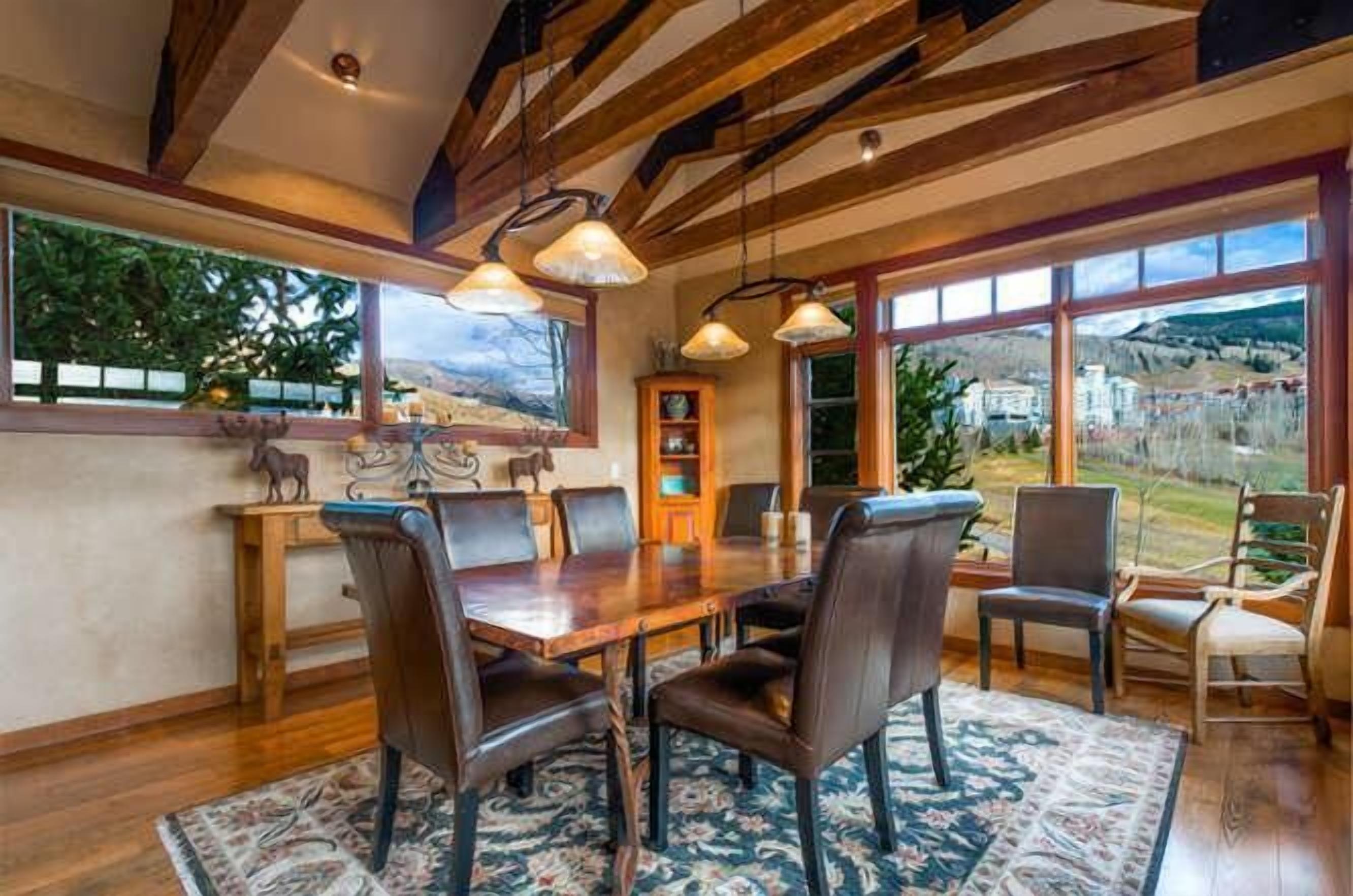 A dining area with a wooden table and chairs, large windows with a view of the outdoors, and a ceiling with exposed beams.