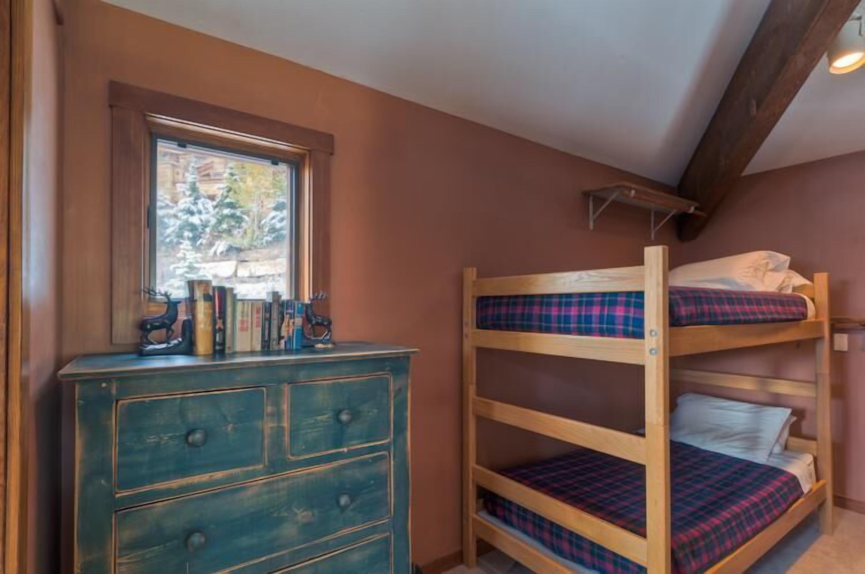 Una cama litera con ropa de cama escocesa, un aparador con cajones y una ventana que da a vistas de árboles.