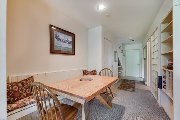 A dining area with a wooden table, chairs, and a bench. There is a framed picture on the wall, a staircase, and a bookshelf with books and decorative items.