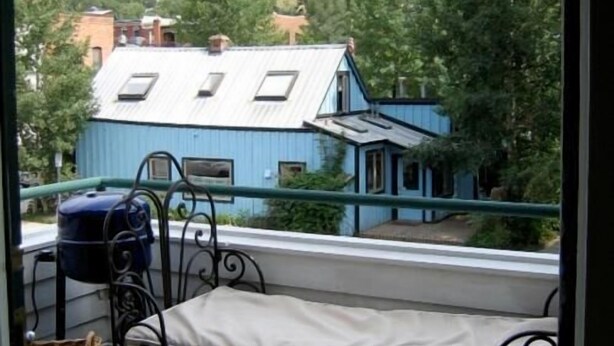 Een balkon met een bed, een stoel en een waterverwarmer. Achtergrond: een blauw huis met een wit dak en een berg.