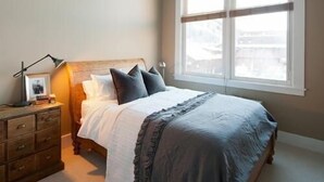 Een slaapkamer met een groot raam, een houten hoofdbord, een nachtkastje met een lamp en een dressoir.