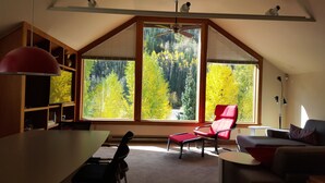 A spacious room with a large window offering a view of a forest. There is a dining table with chairs, a red chair, and a sofa. - Plunge H 3 Bedroom Condo By Accommodations in Telluride (Telluride)