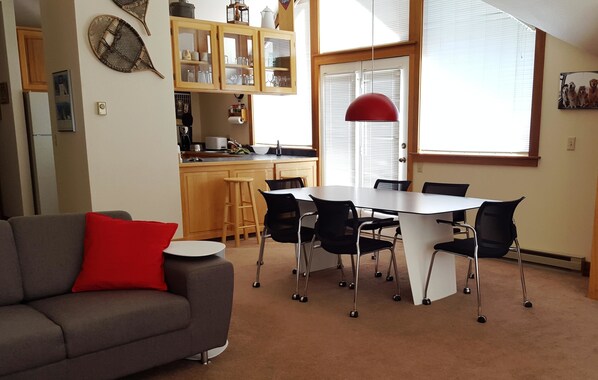 A living room with a gray sofa and a red pillow, a dining area with a white table and black chairs, and a kitchen with wooden cabinets and a red pendant light.