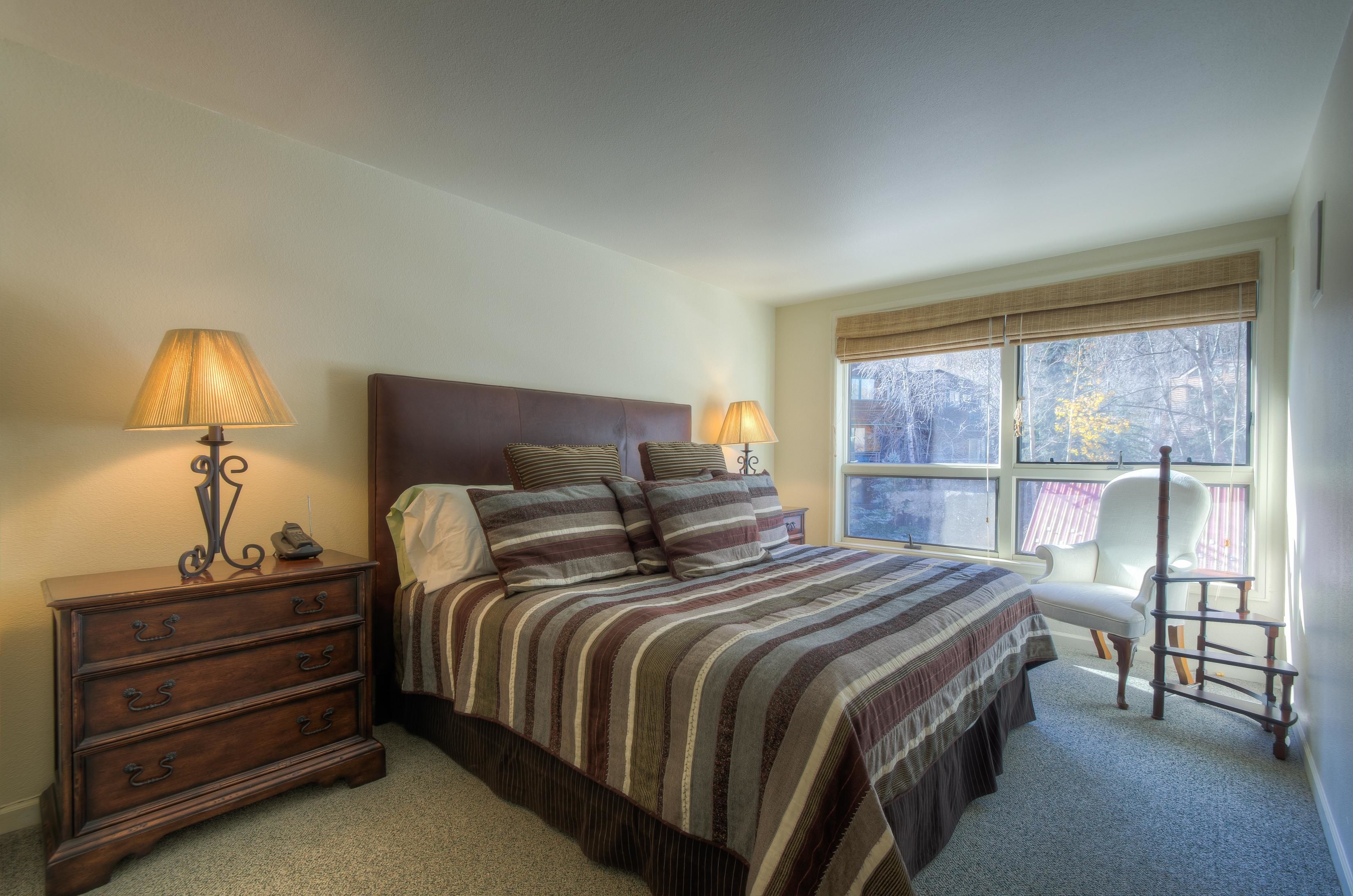 A bedroom with a large bed, two bedside lamps, a wooden dresser, and a chair by the window.