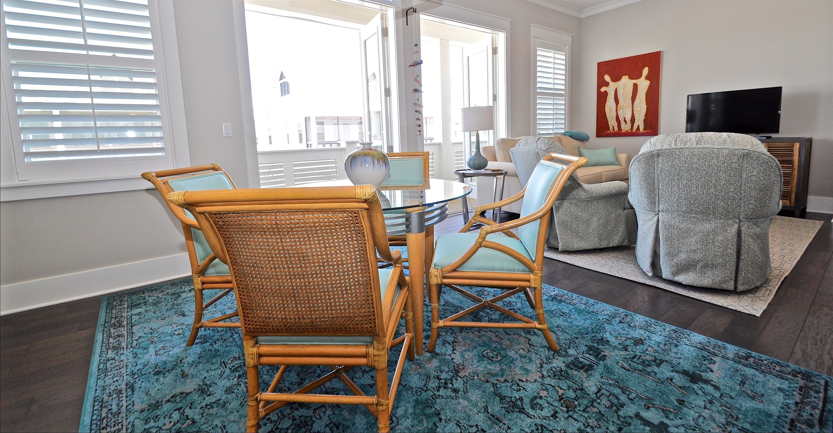 A living room with a dining table, two wicker chairs, a grey armchair, a blue patterned rug, and a TV on a wooden stand.