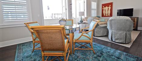 A living room with a dining table, two wicker chairs, a grey armchair, a blue patterned rug, and a TV on a wooden stand.