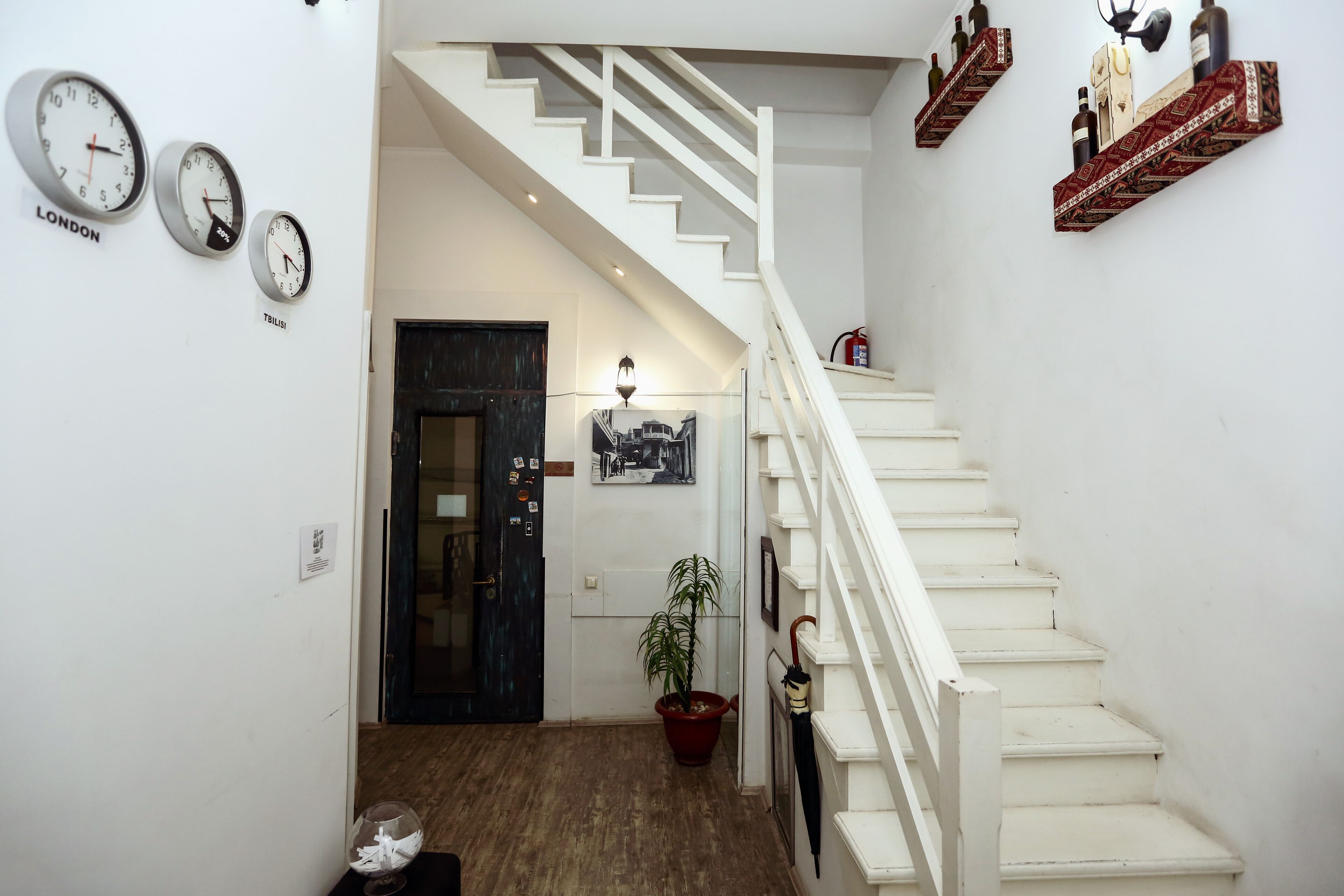 A staircase with white railings leading to a landing. There are clocks on the wall, a potted plant, and a door with a black frame.