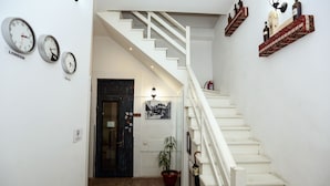 Una escalera con barandillas blancas que conduce a una plataforma. Hay relojes en la pared, una planta en maceta y una puerta con marco negro.