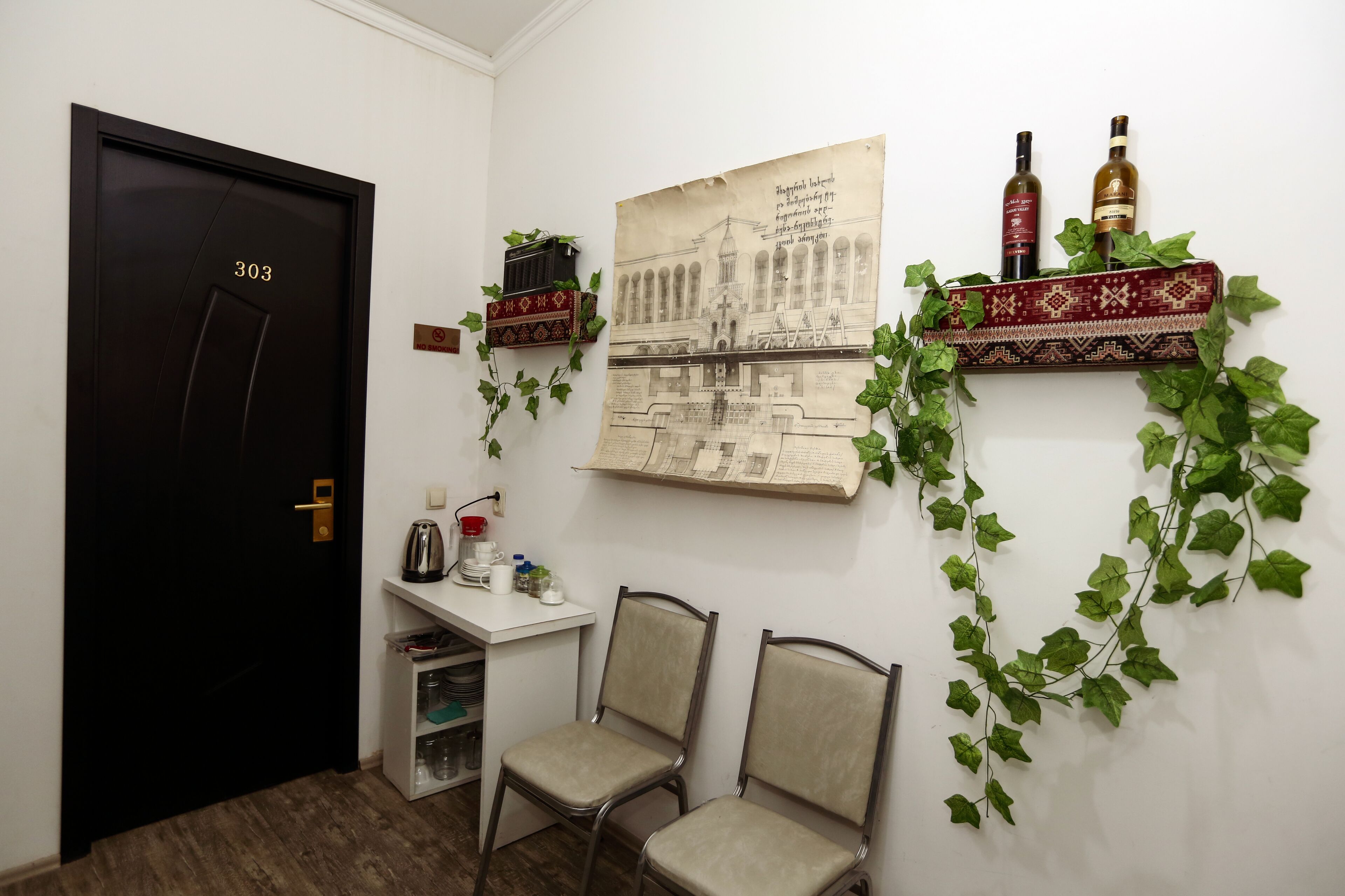 Ein Zimmer mit einer schwarzen Tür mit der Aufschrift 303, einem Schreibtisch mit Wasserkocher, zwei Stühlen, einem Regal mit zwei Flaschen und einem an der Wand hängenden Bild.