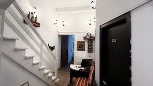 Una escalera que lleva al segundo piso, una puerta negra con la etiqueta "201", una mesita con una silla y un estante en la pared con botellas.