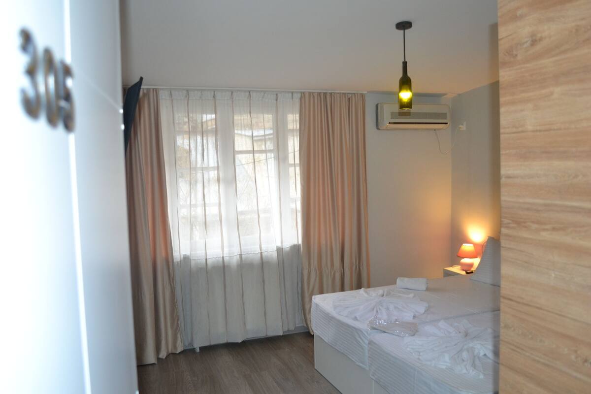 A hotel room with a bed, curtains, an air conditioning unit, a lamp, and a wooden wall.