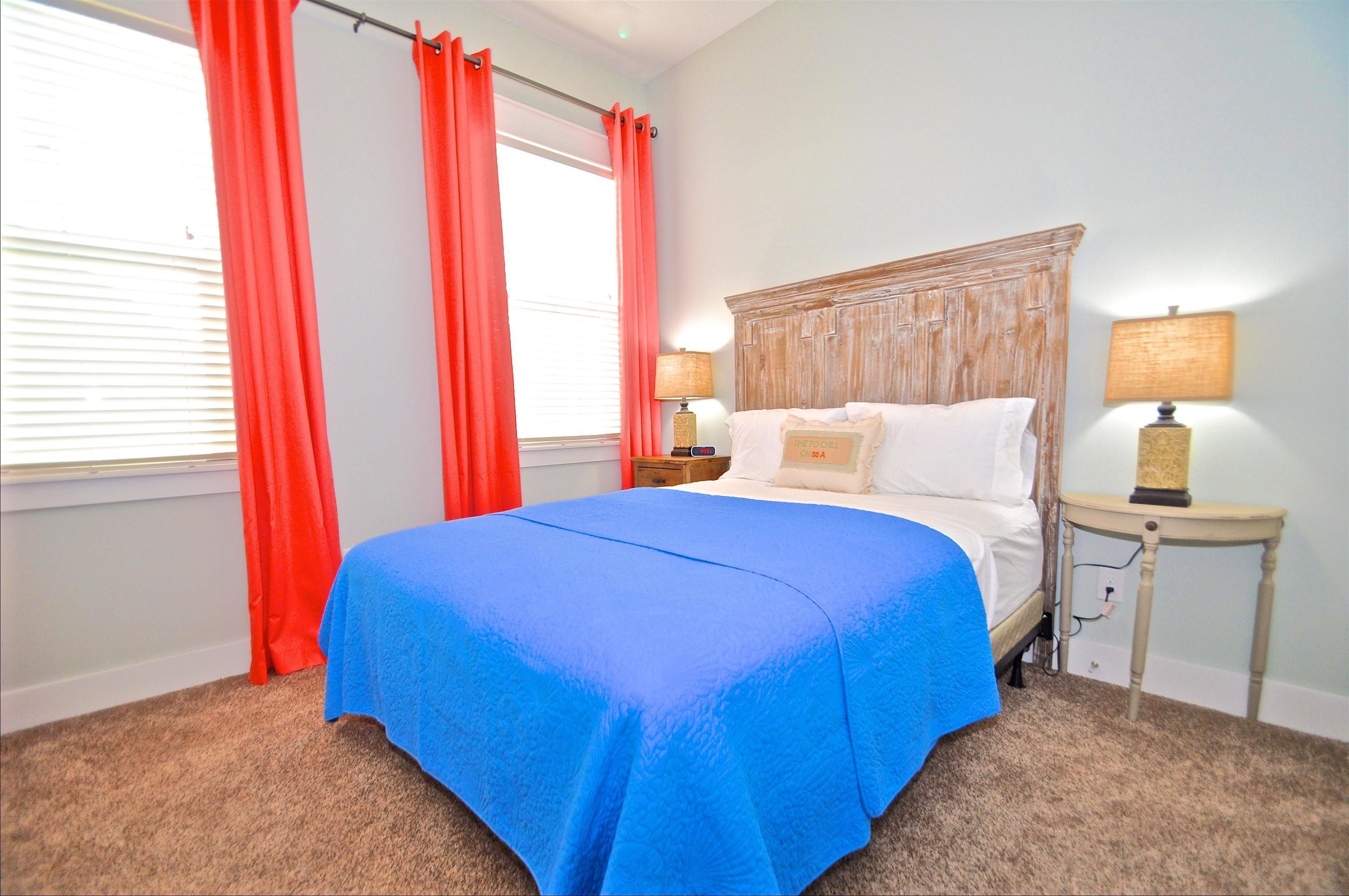 A bedroom with a large bed, two bedside tables, a window with red curtains, and a lamp on each table.