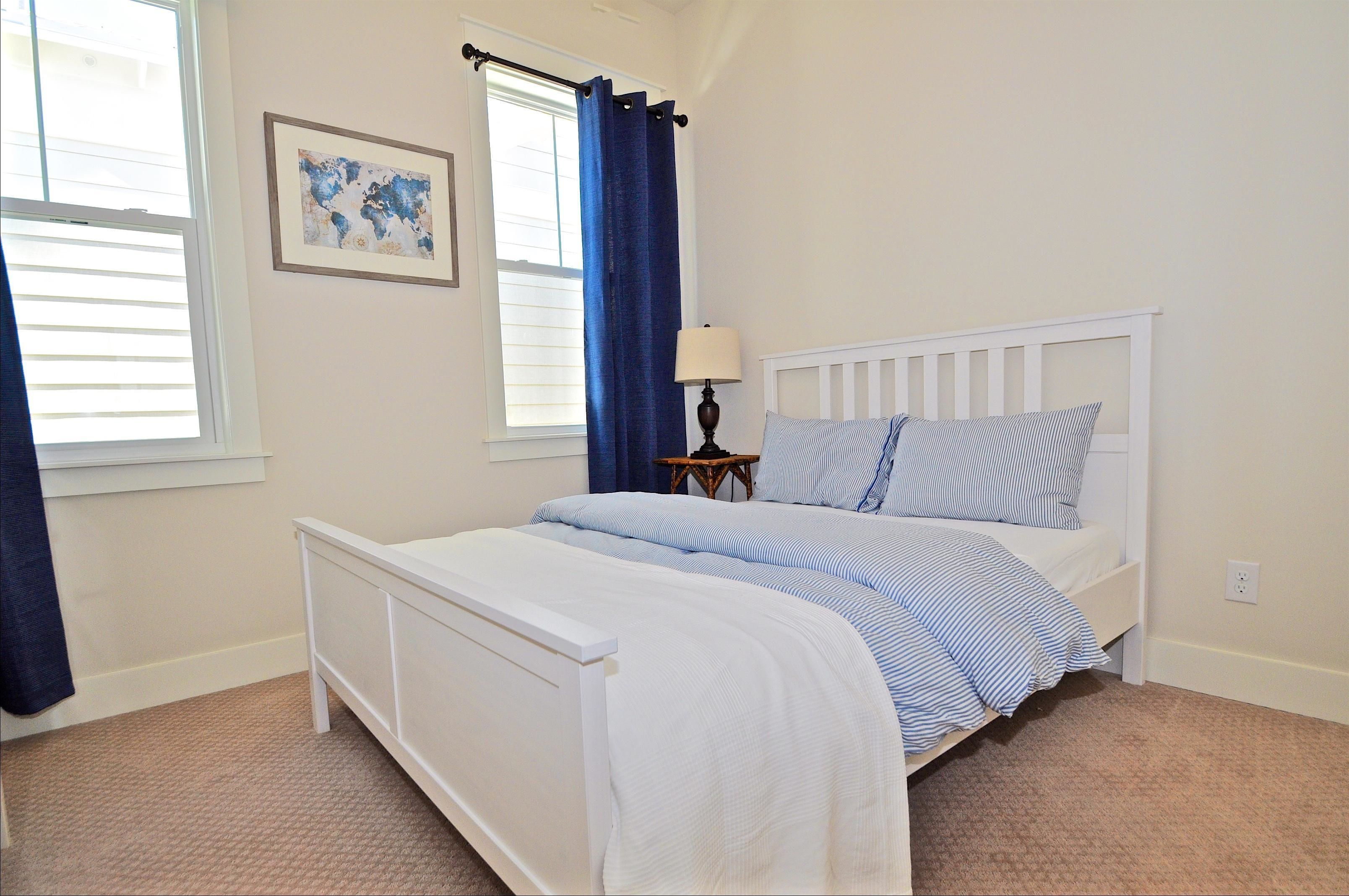 A bedroom with a white bed, blue and white bedding, a small bedside table with a lamp, and a window with blue curtains.