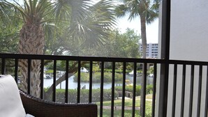 A balcony with wicker furniture, a view of a water body, palm trees, and a building in the background. - Cove II 822F 2 Bedroom Condo By Midnight Cove II (Siesta Key)