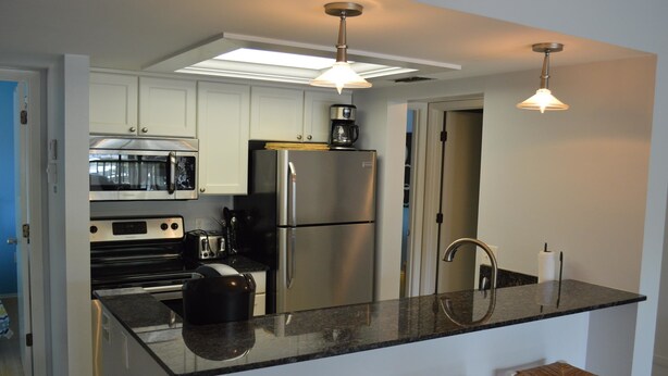 A modern kitchen with stainless steel appliances, a black countertop, and a bar area with stools. - Cove II 822F 2 Bedroom Condo By Midnight Cove II (Siesta Key)