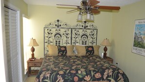 A bedroom with a floral bedspread, a ceiling fan, two bedside lamps, a decorative headboard, and a poster on the wall. - Cove II 832F 2 Bedroom Condo By Midnight Cove II (Siesta Key)