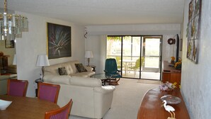 A living room with a sofa, chairs, a dining table, and a view through a glass door to an outdoor area. - Cove II 912C 2 Bedroom Condo By Midnight Cove II (Siesta Key)