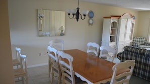 A dining area with a wooden table and white chairs, a chandelier, a mirror, and a cabinet with shelves. - Cove II 922C 2 Bedroom Condo By Midnight Cove II (Siesta Key)