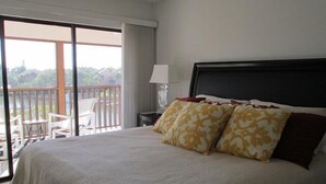 A hotel room with a large bed, two decorative pillows, a nightstand with a lamp, and a view of a balcony with chairs.