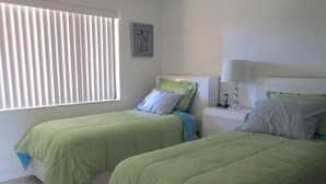 A room with two beds, each with green bedding, a bedside table with a lamp, and a window with blinds.