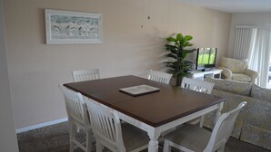 A dining area with a wooden table and white chairs, a television on a side table, and a sofa with floral upholstery. - Cove II 934C 2 Bedroom Condo By Midnight Cove II (Siesta Key)