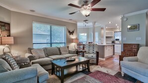 A spacious living room with a sofa set, a coffee table, and a patterned rug. A dining area with chairs and a chandelier is visible in the background.