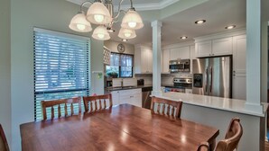 A modern kitchen with stainless steel appliances, a dining area with a wooden table and chairs, and a large window with blinds.