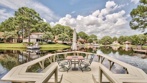 A wooden deck with a table and chairs overlooking a calm lake, surrounded by lush greenery and residential buildings.