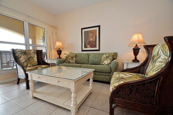 Una sala de estar con una mesa de centro de vidrio, un sofá verde y dos sillones con tapicería floral.