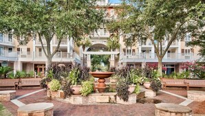 A courtyard with a central fountain, surrounded by potted plants and benches, with a multi-story building in the background.