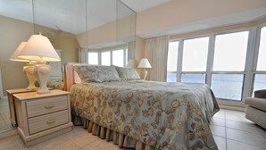 A bedroom with a large bed, two bedside lamps, a dresser, and a view of the sea through large windows.