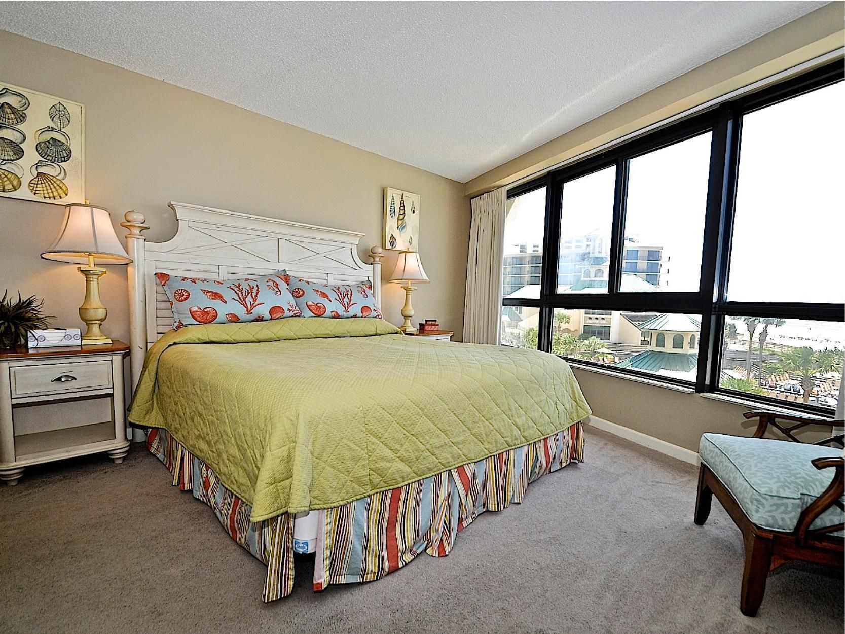 A bedroom with a large bed, two bedside tables, a chair, a window with a city view, and artwork on the walls.