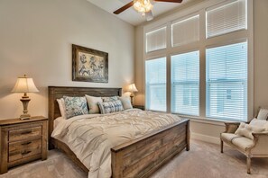 A bedroom with a wooden bed, two bedside tables with lamps, a chair, and large windows with blinds. - JACK's Place 3 Bedroom Condo By Coastal Dreamin (Santa Rosa Beach)