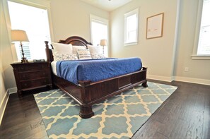 A bedroom with a wooden bed, a nightstand, a lamp, and a blue patterned rug. - Sandlot 3 Bedroom Condo By Coastal Dreamin (Santa Rosa Beach)