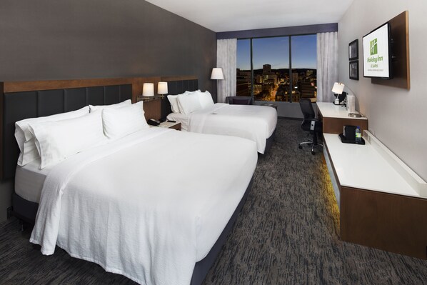 Habitación de hotel con dos camas, un escritorio y un ventanal grande con vistas a la ciudad.