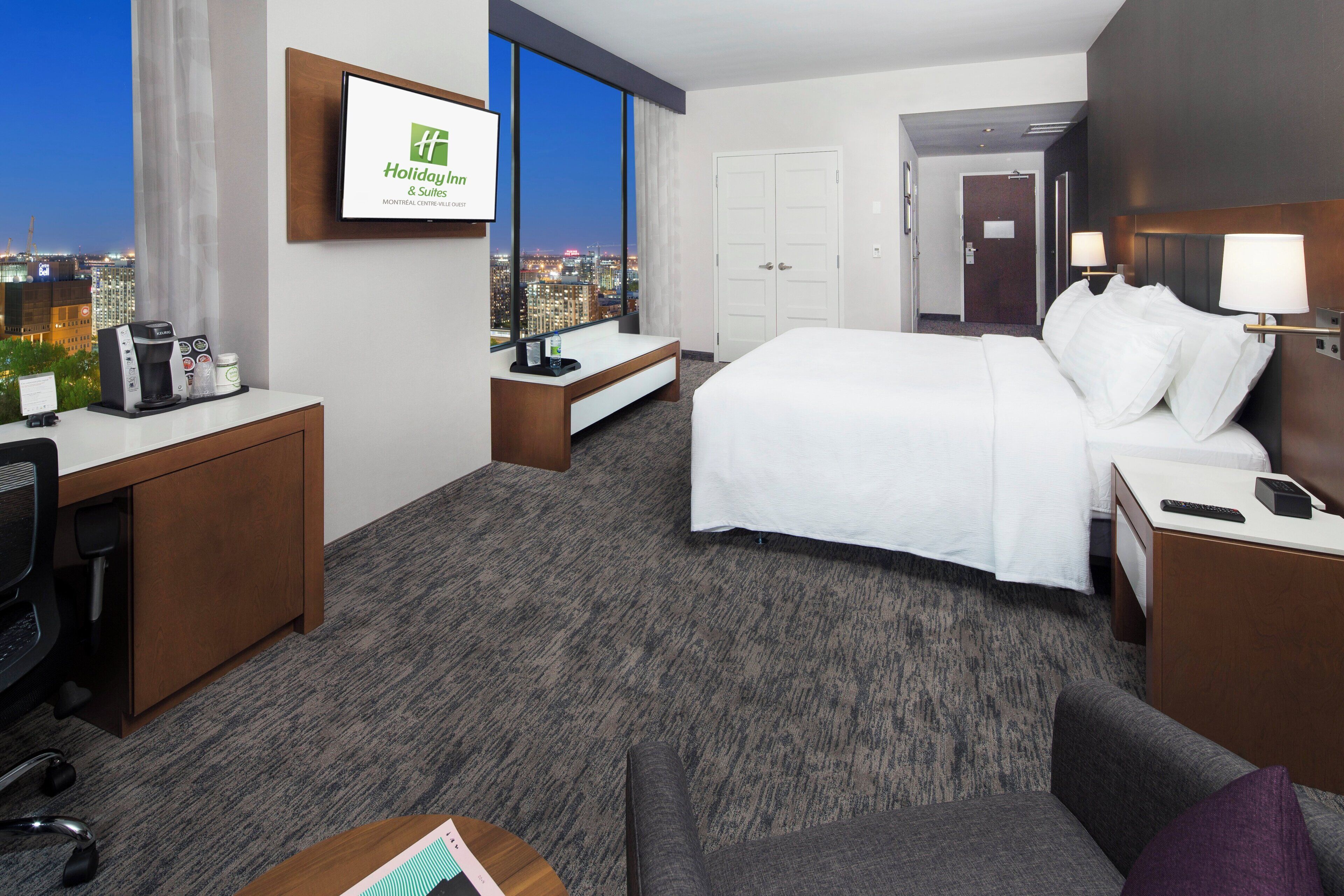 A hotel room with a bed, a desk, a chair, a sofa, a coffee maker, and a view of the city at night.