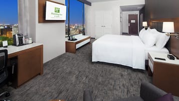 A hotel room with a bed, a desk, a chair, a sofa, a coffee maker, and a view of the city at night.