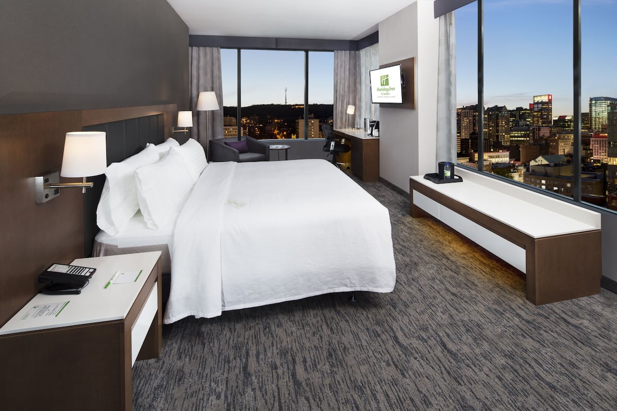 A modern hotel room with a large bed, a desk, and a view of the cityscape.