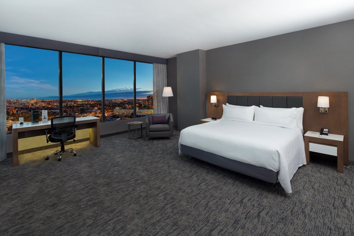 A hotel room with a large bed, a desk, a chair, and a view of a cityscape through large windows.