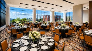 A spacious banquet hall with round tables set for dining, large windows offering a city view, and a modern ceiling design.
