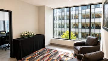 Un salón de conferencias con una mesa negra, flores blancas, una silla de cuero marrón, alfombra estampada y un amplio ventanal con vista a un edificio.