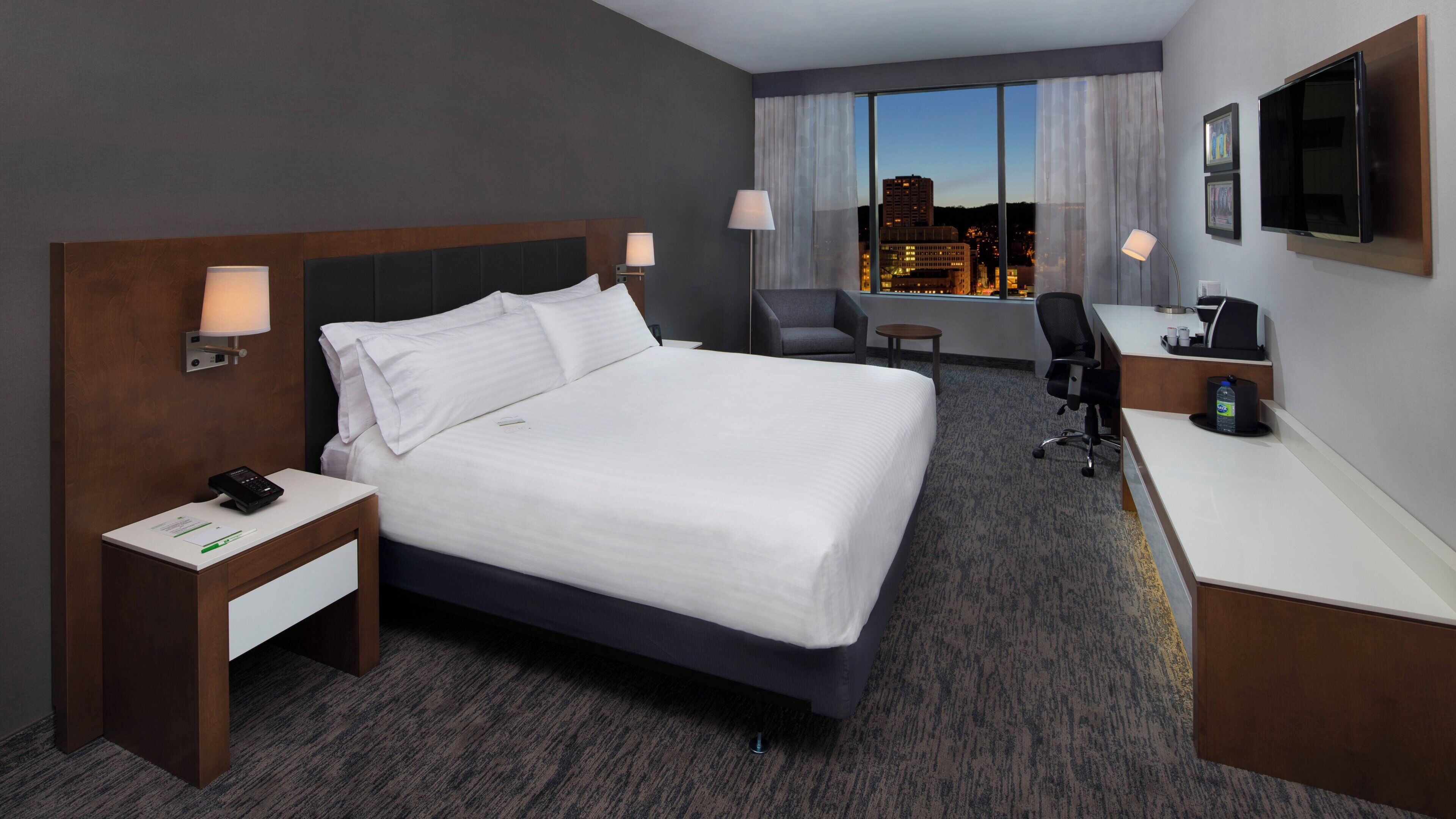A hotel room with a bed, desk, chair, and a view of the cityscape.