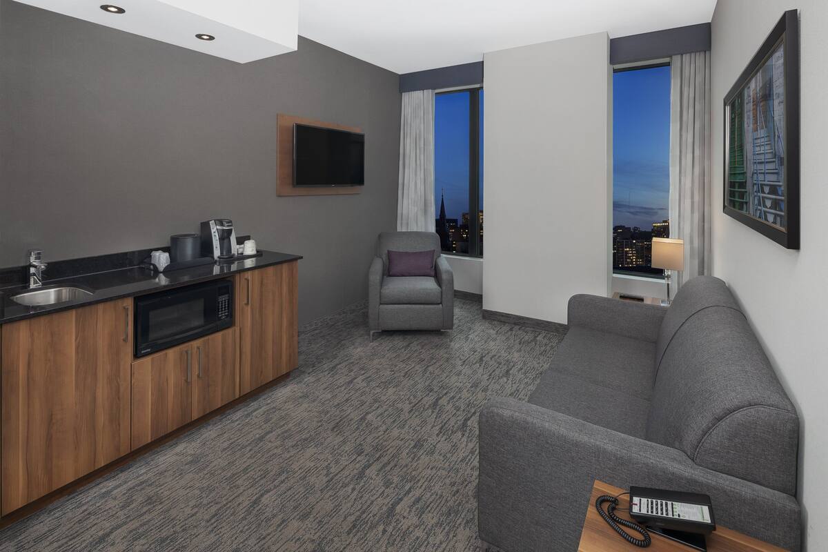 A modern hotel room with a kitchenette, grey sofa, and a city view through the window.