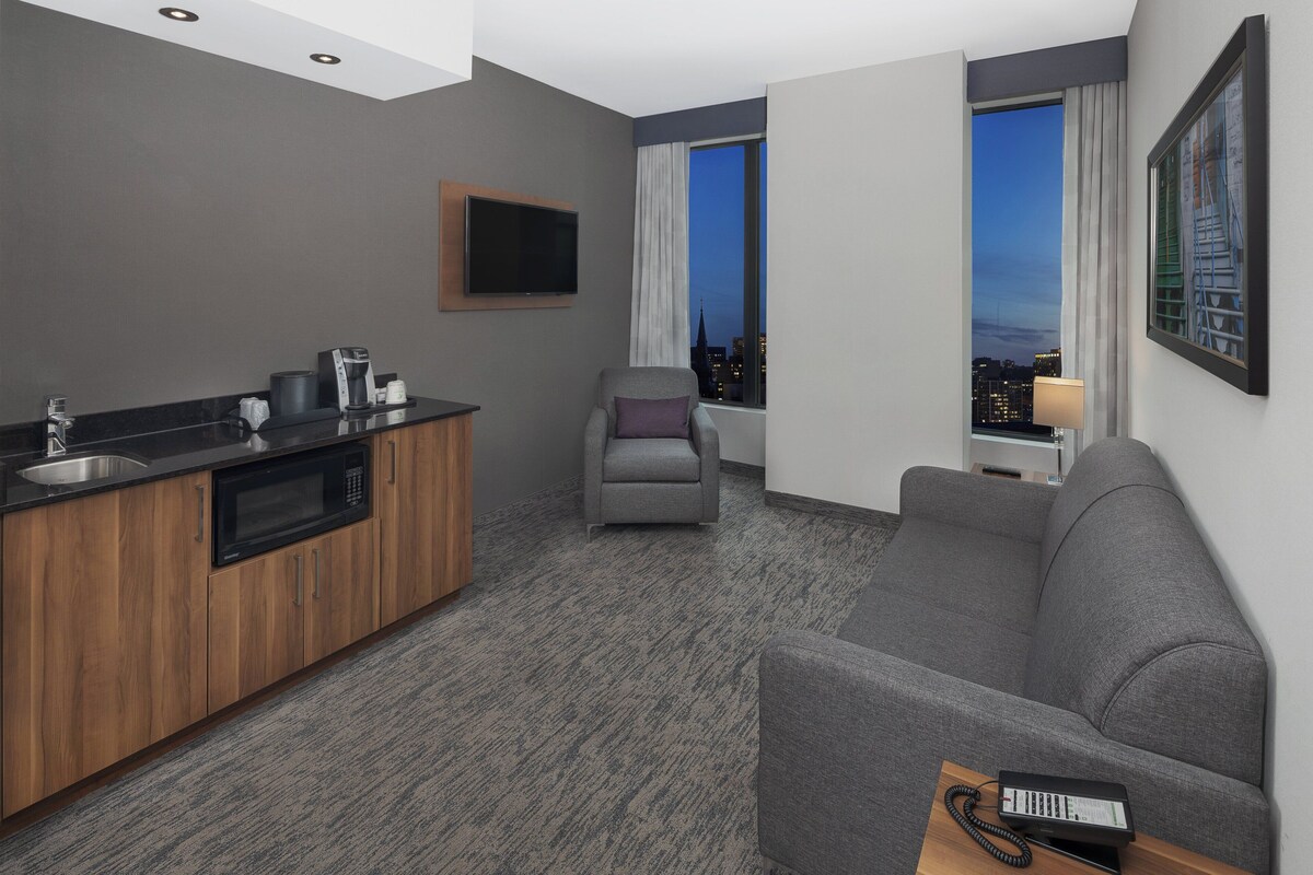 A modern hotel room with a kitchenette, grey sofa, and a city view through the window.