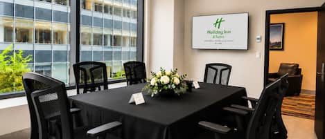 Une salle de conférence avec une grande table, des chaises noires, un arrangement floral au centre et un écran affichant le logo de « Holiday Inn & Suites ».