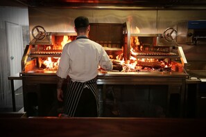 A chef in a white shirt and striped apron cooking in a professional kitchen with a large open flame grill.