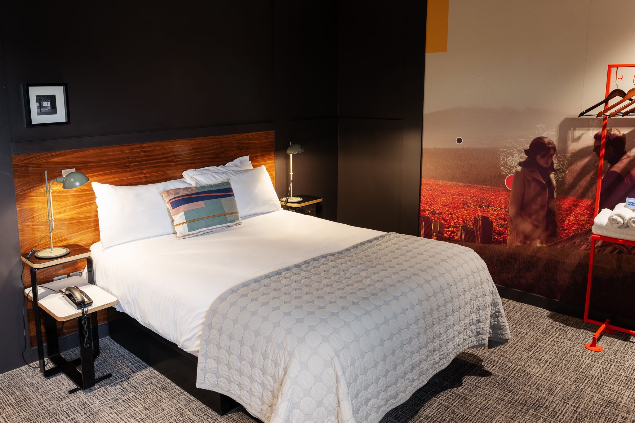 A hotel room with a bed, bedside tables, a lamp, a phone, and a poster on the wall.