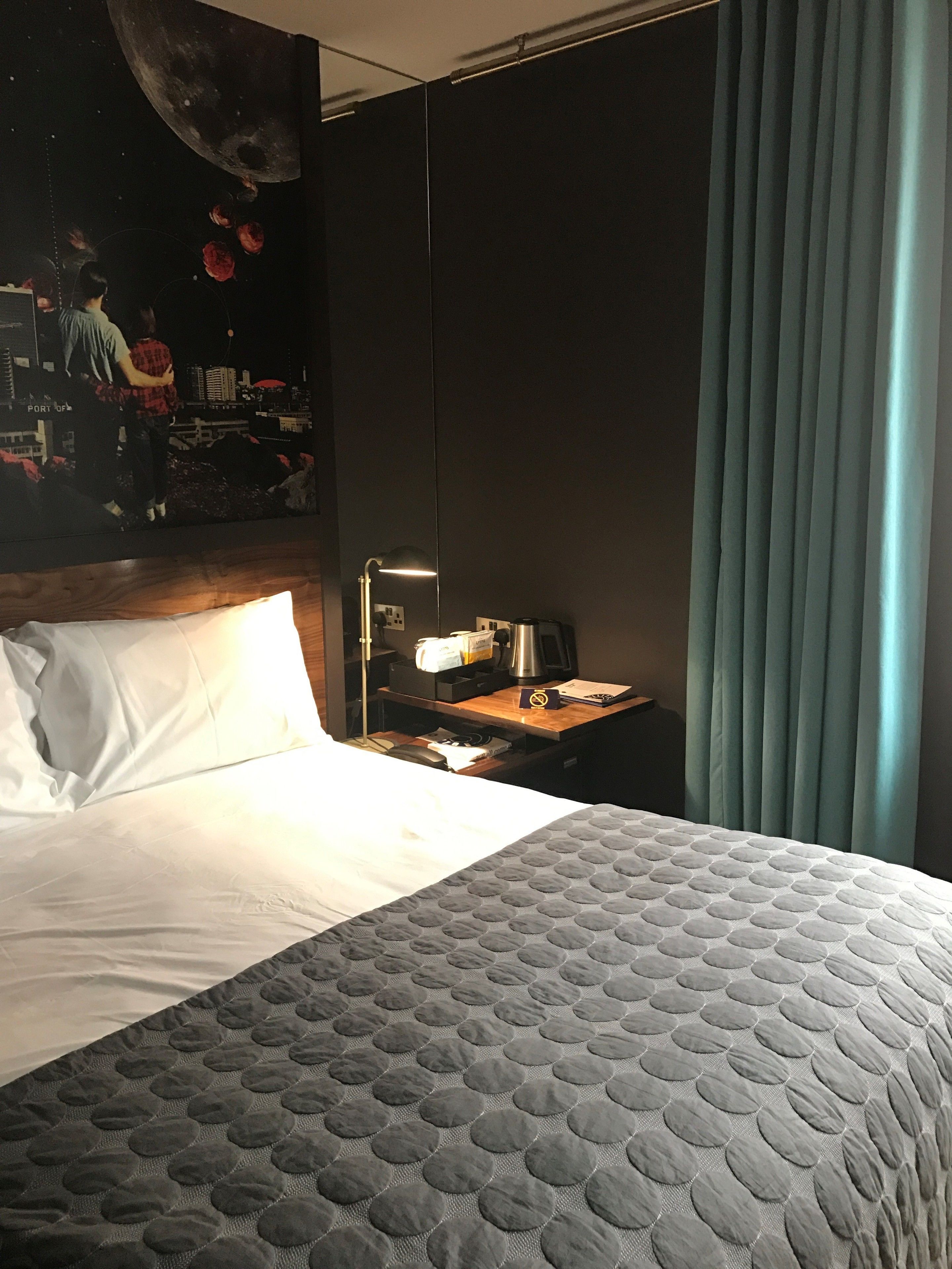 A hotel room with a bed, a nightstand with a lamp, a painting on the wall, and a window with curtains.